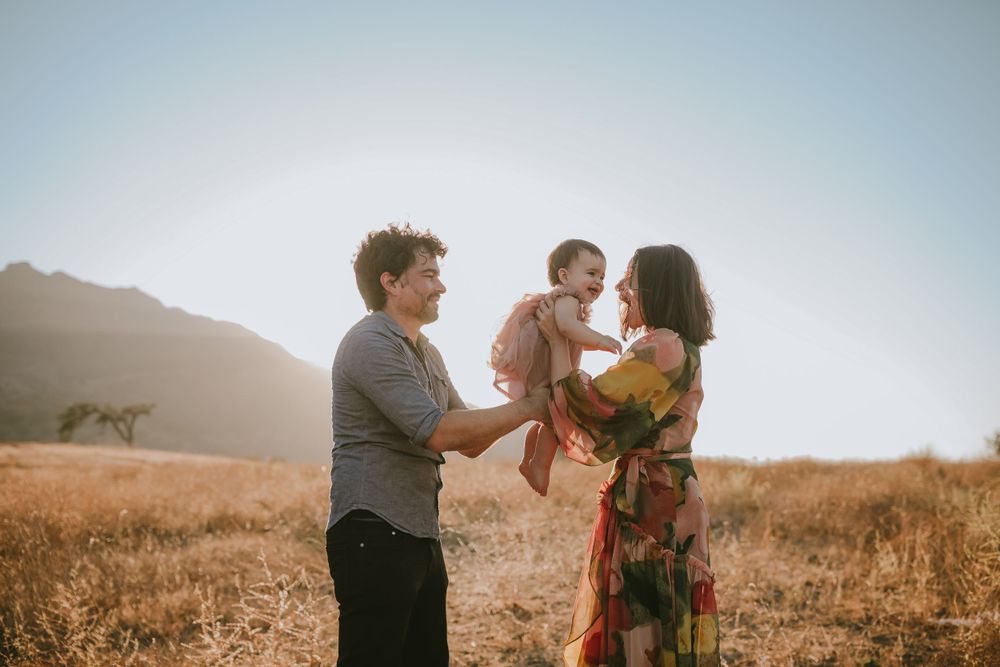 Family shares tender moment together in golden field during sunset with mountains in background.