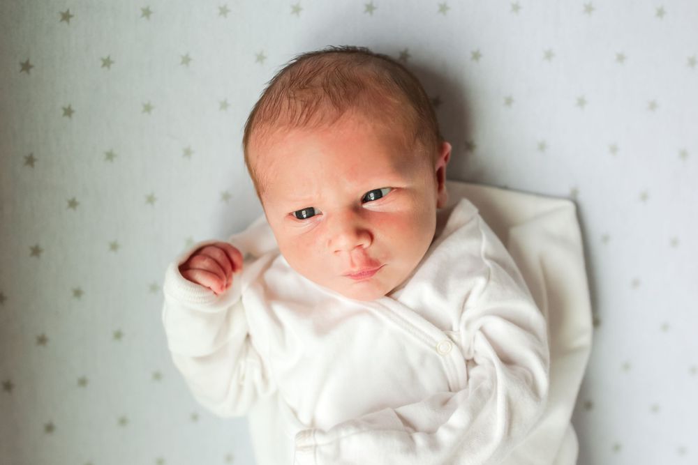 A newborn baby wearing white clothing lies on a polka dot sheet while looking upward.