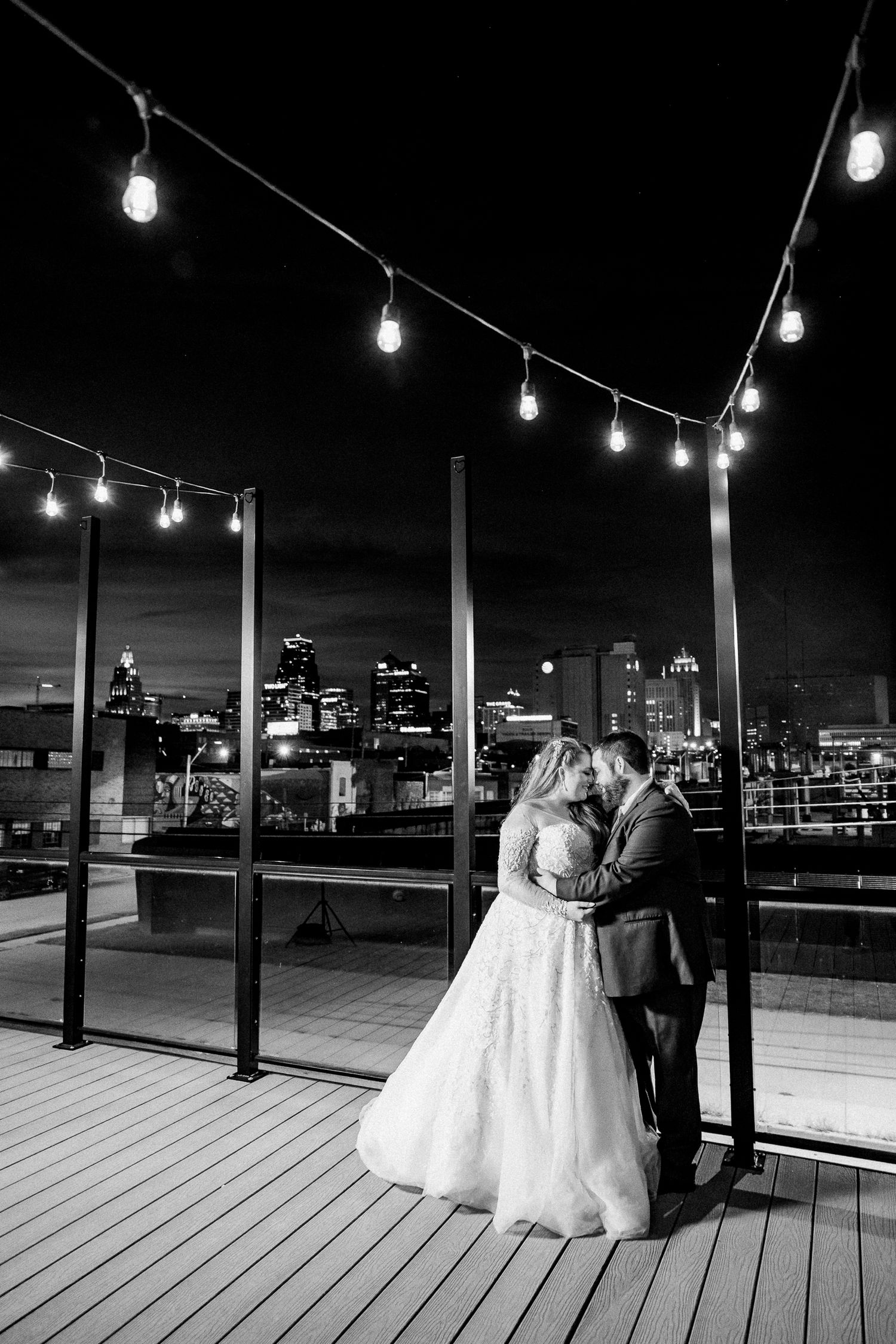 A romantic wedding couple embraces on a rooftop deck at night with string lights and city skyline in the background by Stephanie Coulter Pho