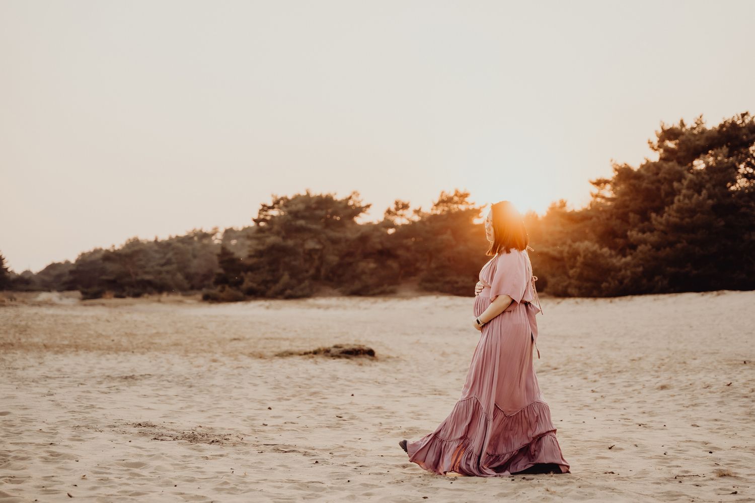 Figure in flowing pink dress stands on beach at sunset with golden sunlight filtering through trees.
