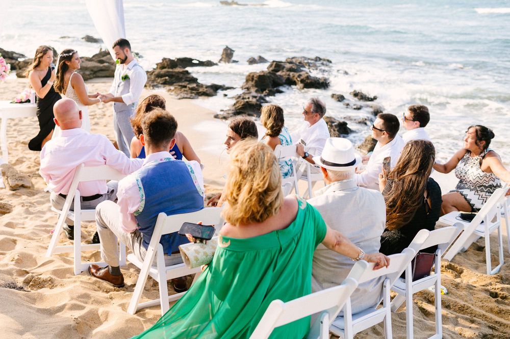 Beach wedding ceremony with guests seated on white chairs overlooking ocean.