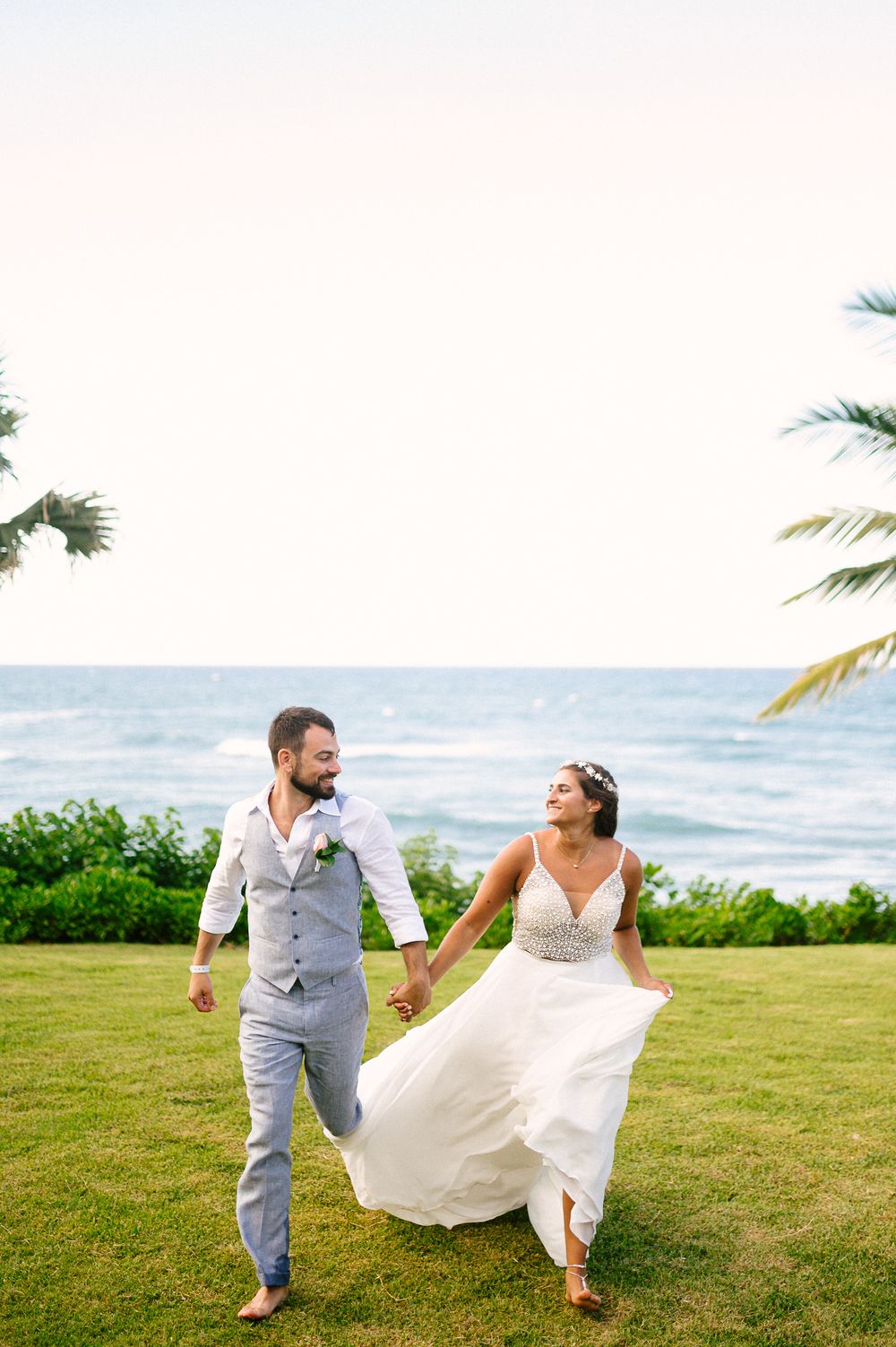 A couple running playfully on oceanfront grass with palm trees swaying overhead.