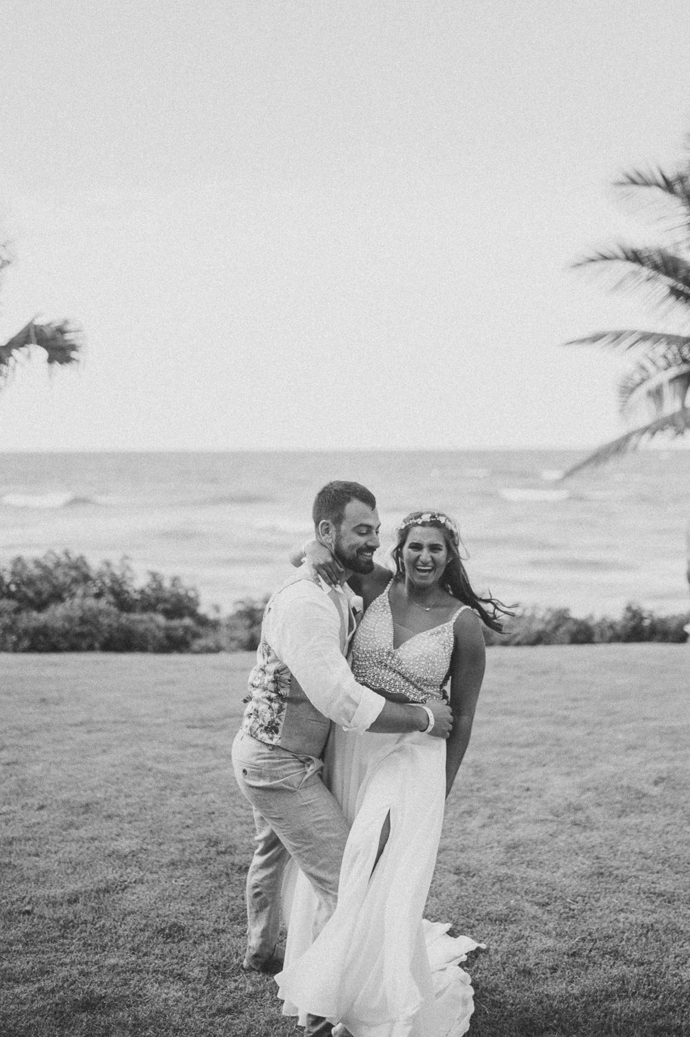A couple shares a joyful moment together on a grassy oceanfront lawn with palm trees in the background.