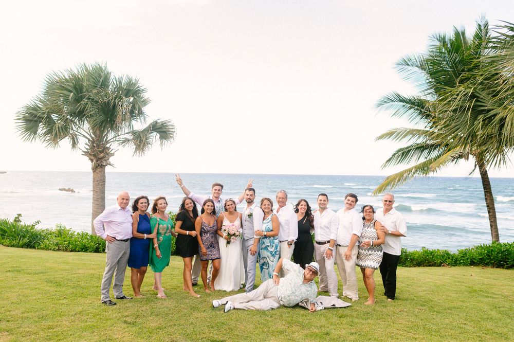 Wedding party gathers for group photo on tropical beach lawn with palm trees and ocean views.