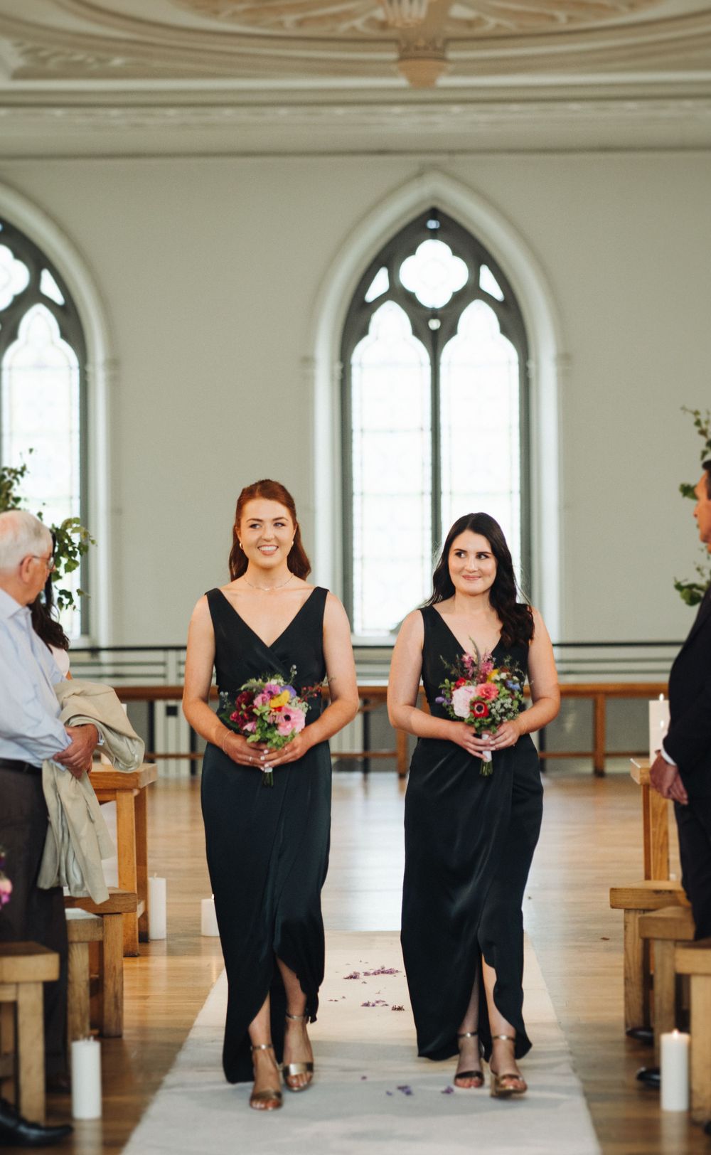 Bridesmaids in elegant black gowns walking down aisle in church with gothic windows.