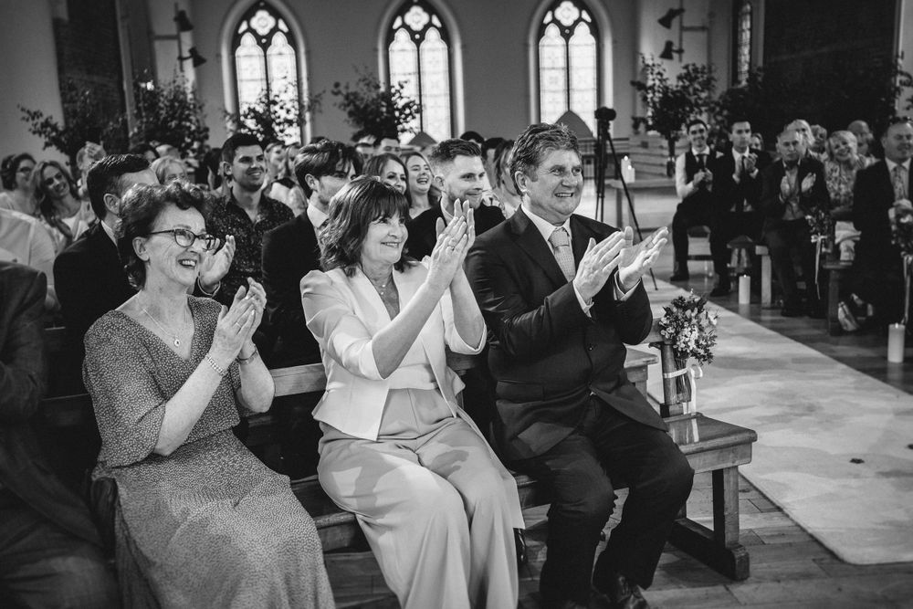 Wedding guests clapping and celebrating during ceremony in historic church with stained glass windows.