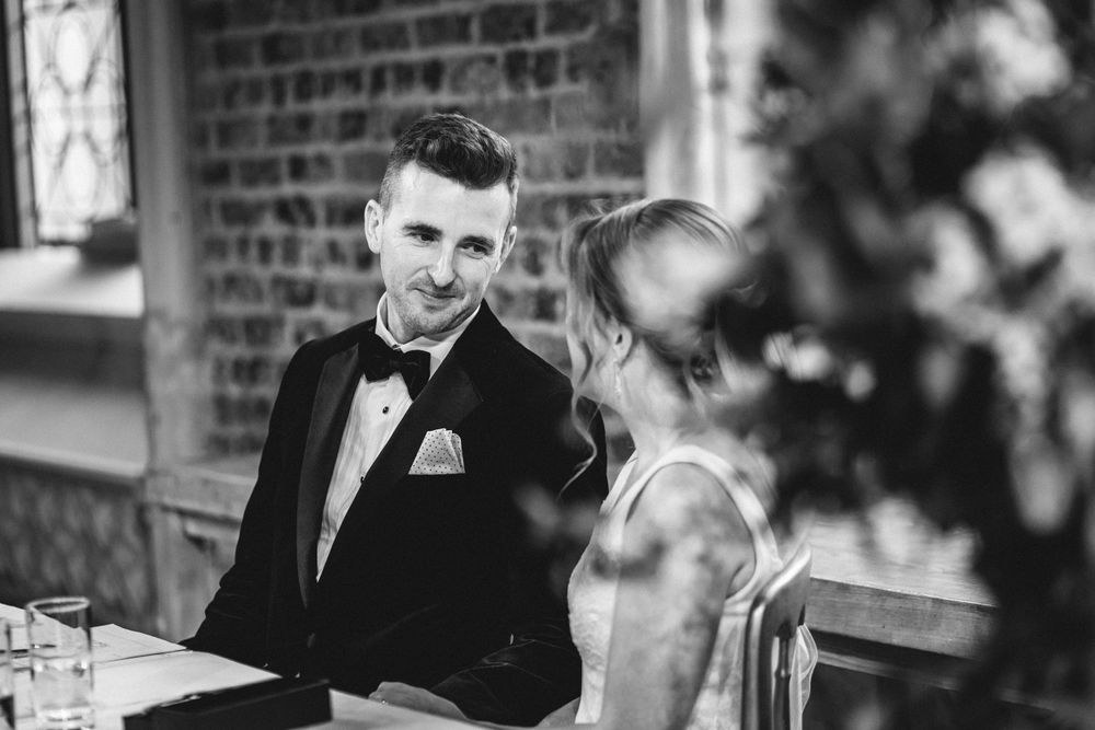 A black and white candid moment of a couple sharing smiles at an indoor wedding reception.