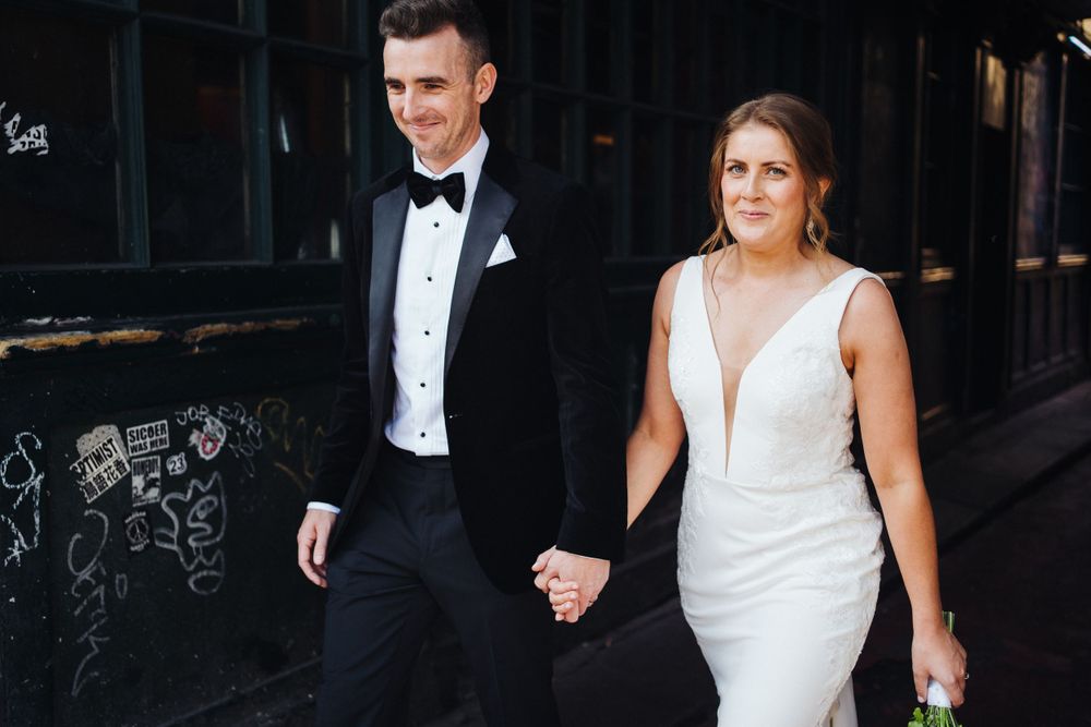Couple in formal attire walking hand in hand past urban graffiti wall.