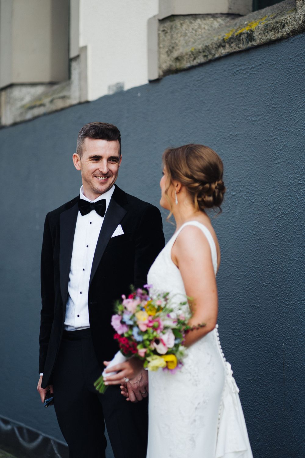 Wedding couple sharing a tender moment against a gray wall with the bride holding a vibrant floral bouquet.