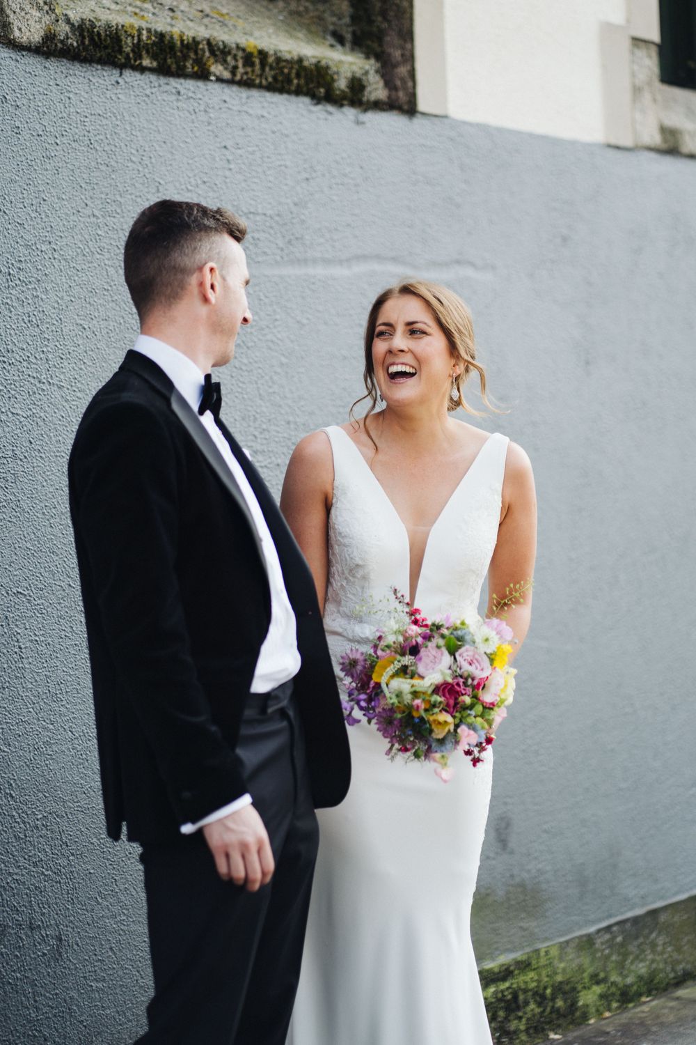 Bride and groom share laughter during their wedding photoshoot against an urban wall backdrop.