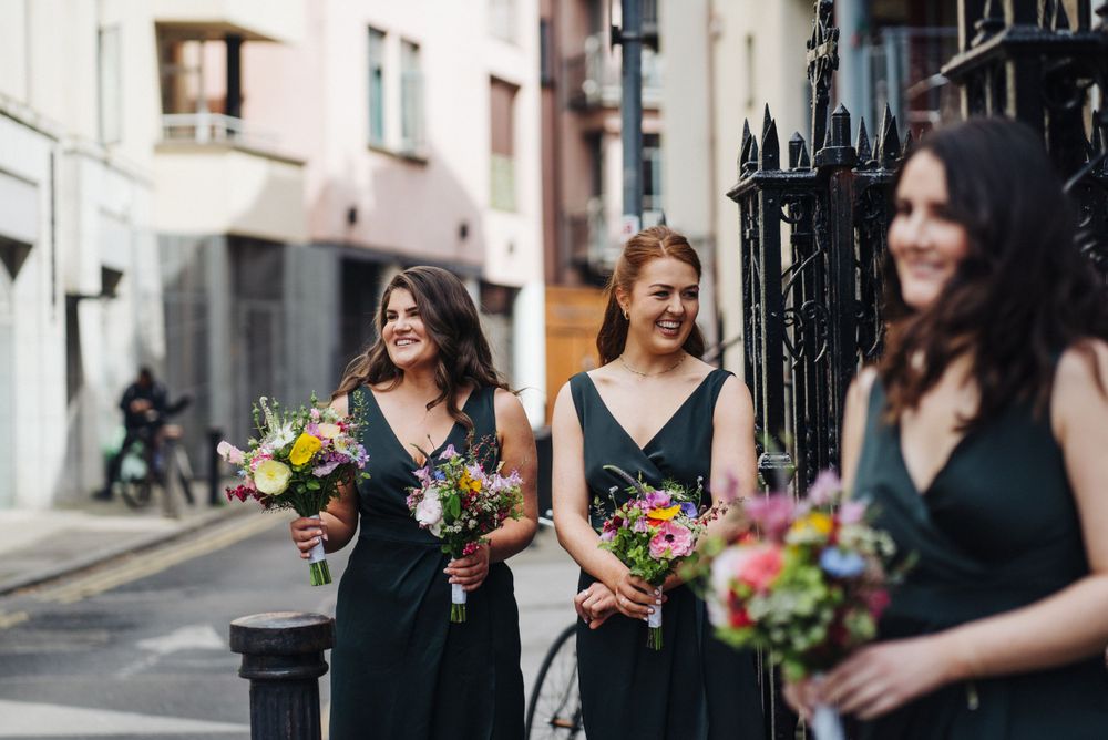 Three bridesmaids in black dresses walking down a city street carrying colorful floral bouquets.