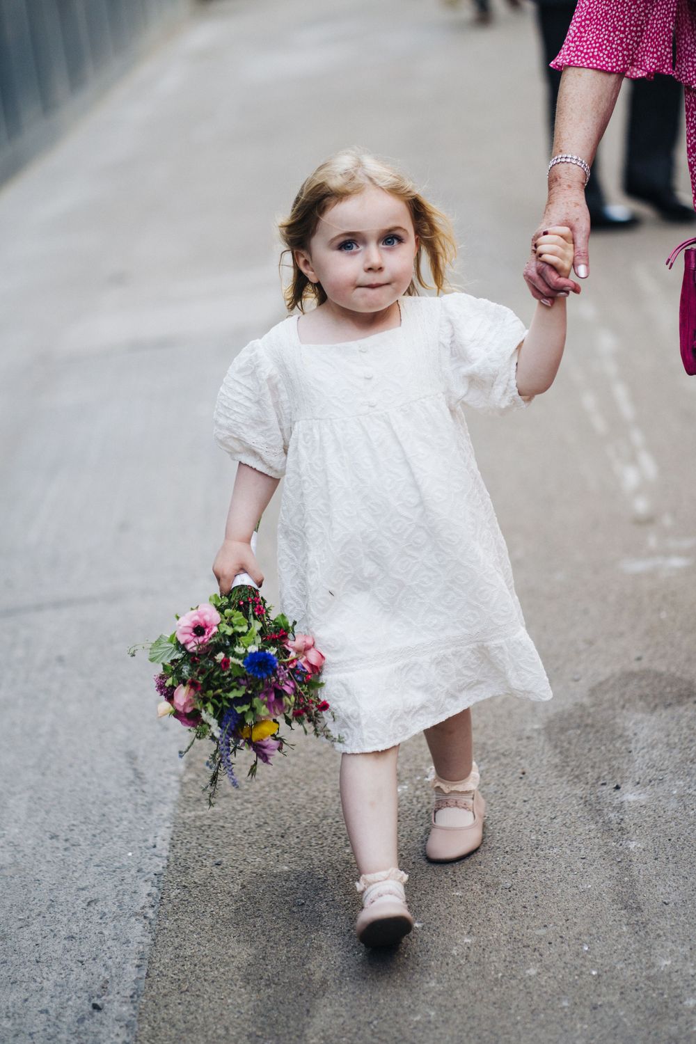 Young child in white dress holding colorful bouquet walks down street.