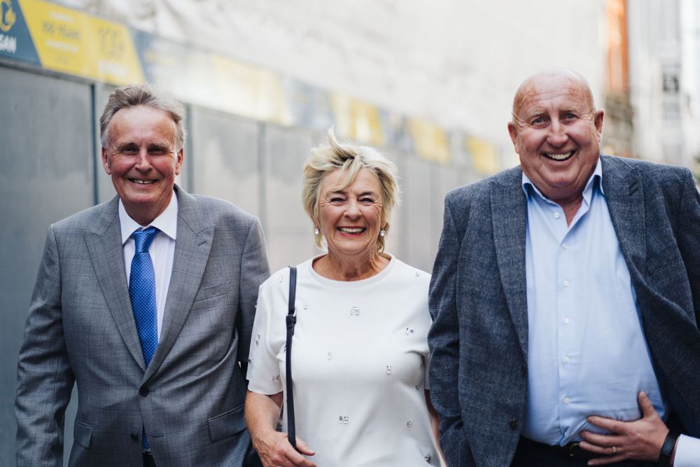 Three elegantly dressed guests in formal attire smiling outdoors.
