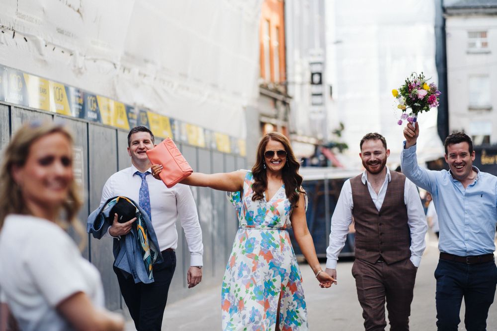 Wedding party members walk together down alley holding hands and bouquets.