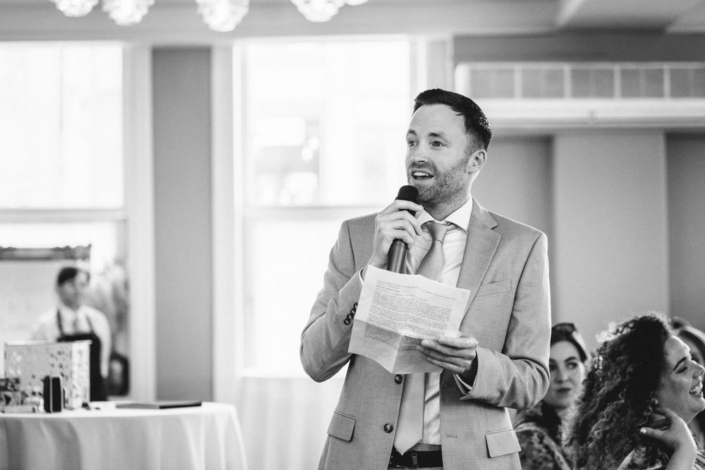 Person in grey suit delivers wedding speech at elegant reception venue.