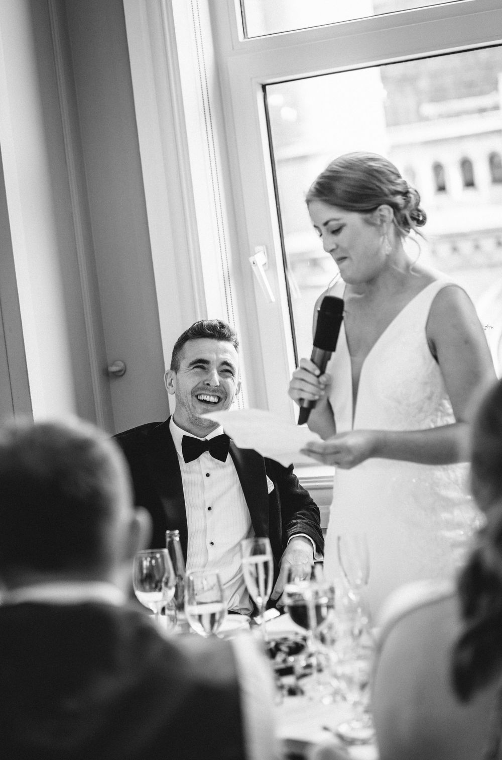 Candid moment captured during wedding reception speeches with elegant window backdrop.