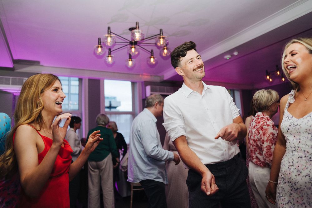 Wedding guests dancing and celebrating under purple lighting and a modern chandelier at an indoor reception.