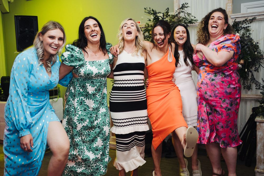Group of friends in colorful dresses laughing and posing together against a bright wall.