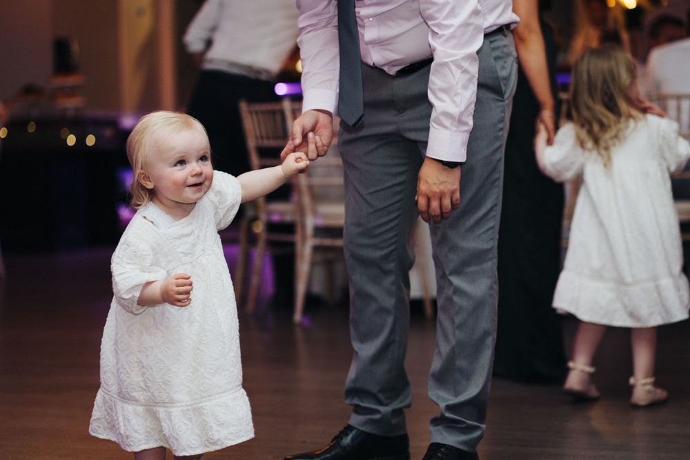 A small child in a white dress dances on the reception floor while holding an adult's hand.