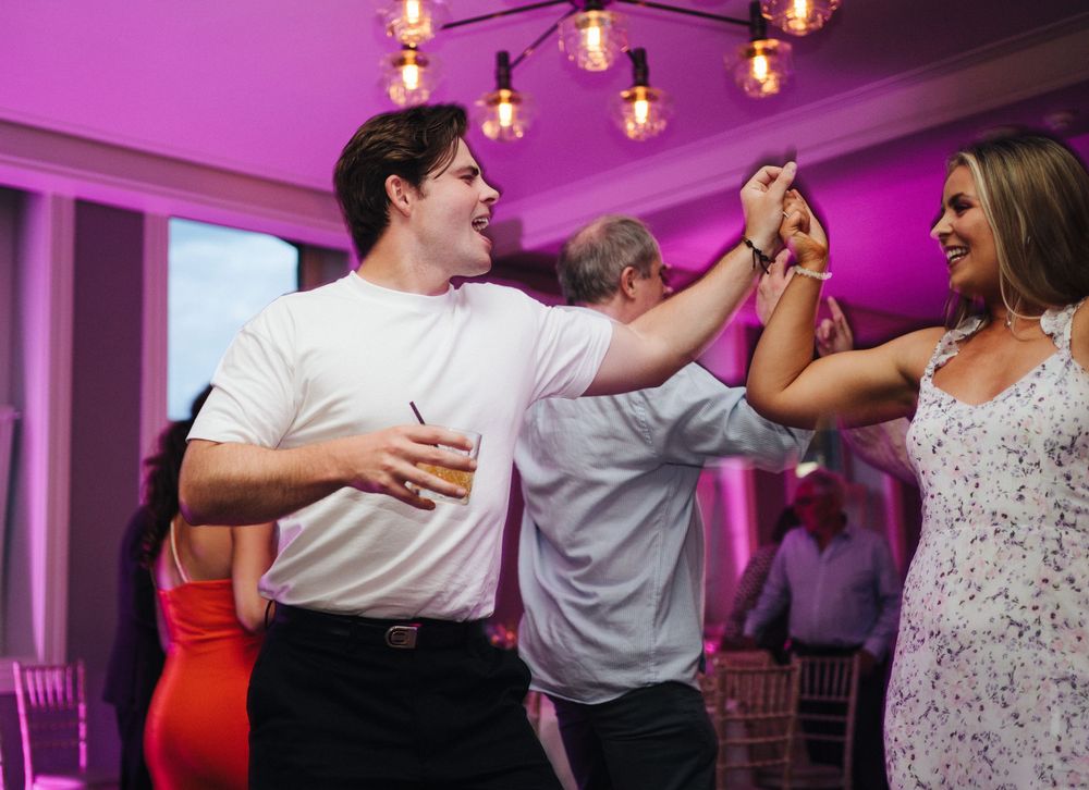 Party guests celebrate and dance with joyful expressions under purple mood lighting at an event.