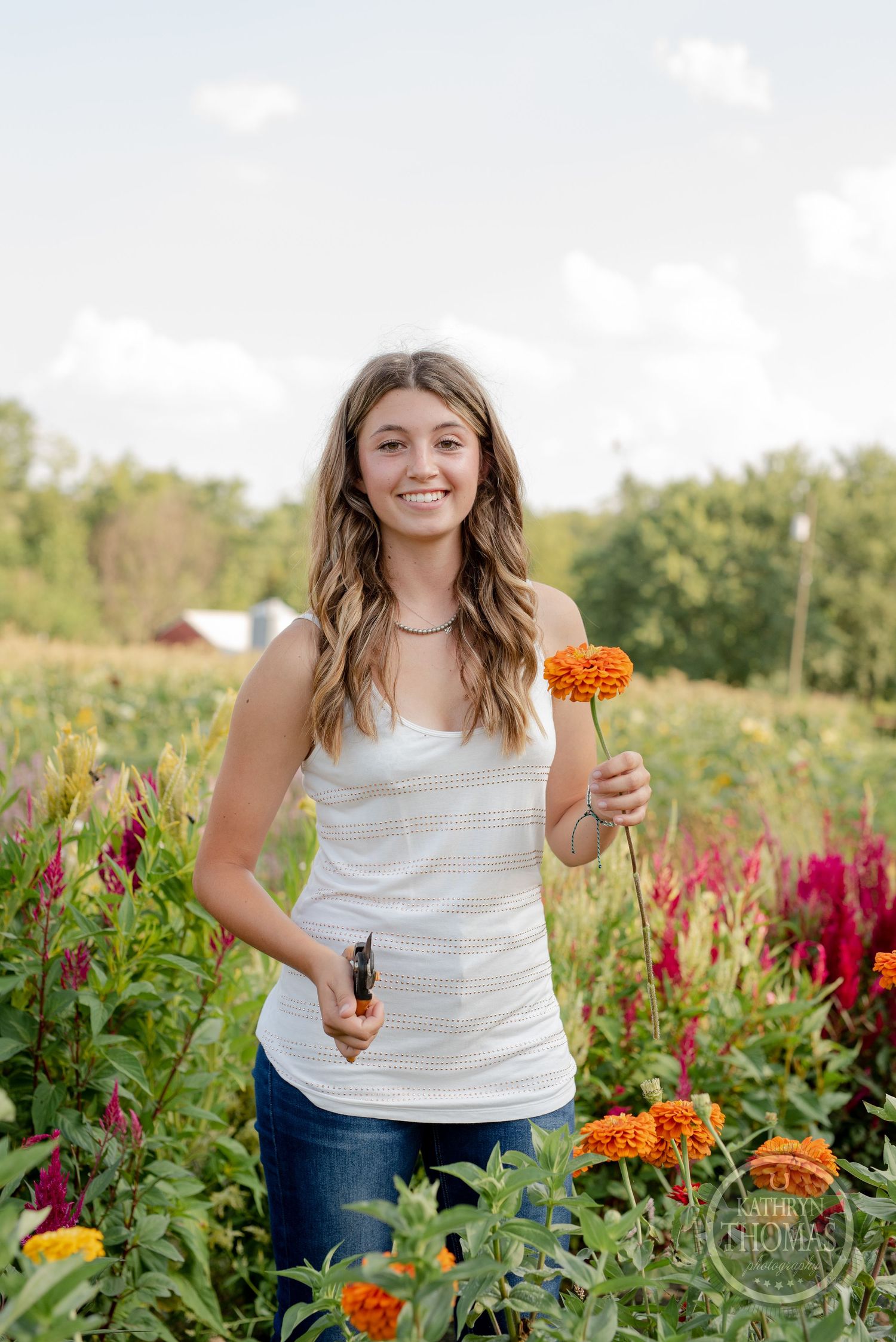 A person holds garden tools and an orange flower while working in a vibrant flower garden.