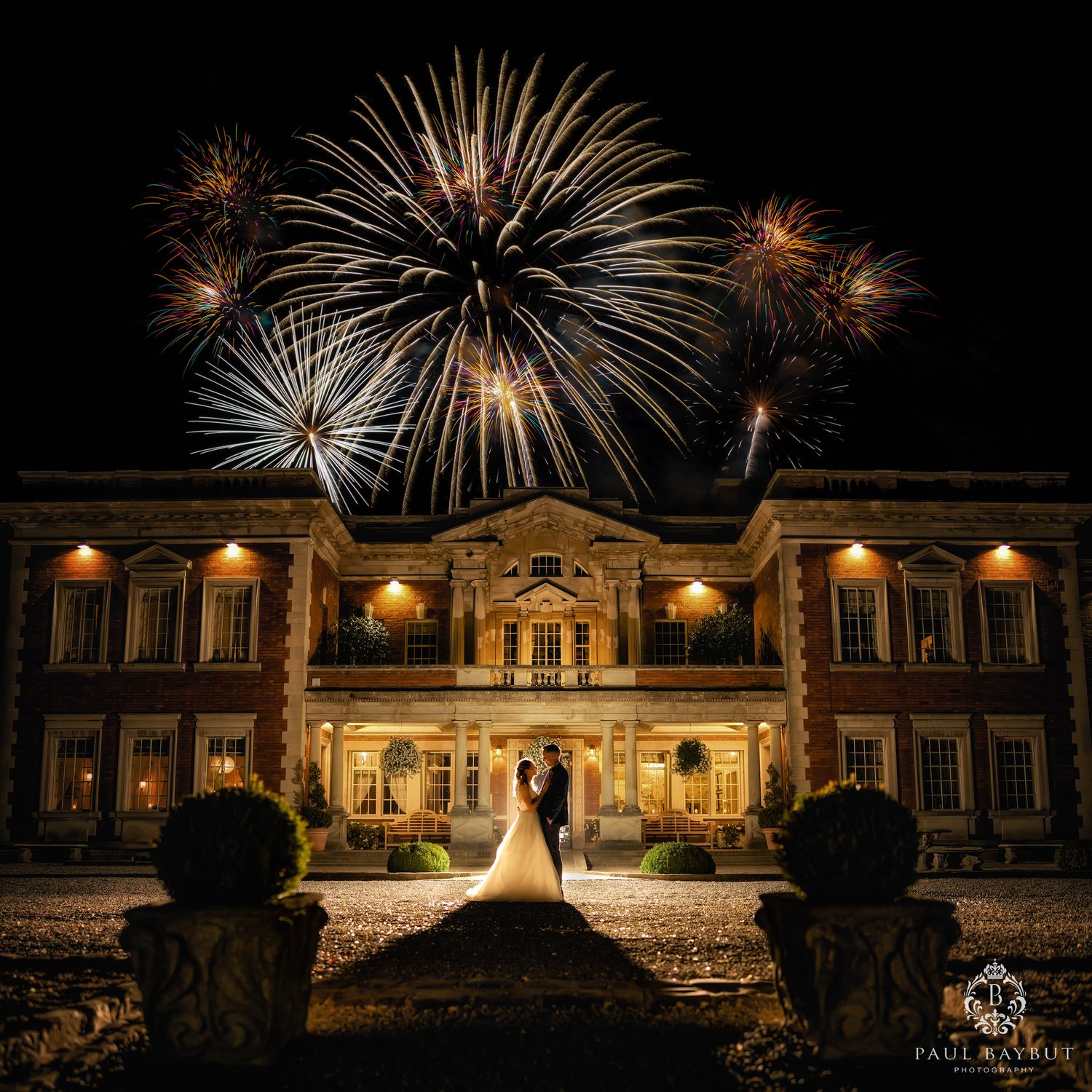 Spectacular fireworks display lights up the night sky above an illuminated historic mansion during a wedding celebration.