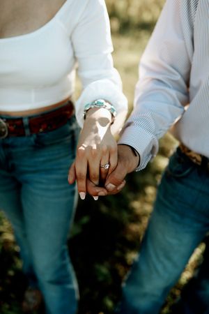 Close up of intertwined hands showing engagement ring in sunlit outdoor setting.