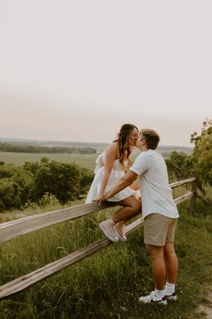 Couple sitting on a wooden fence at sunset overlooking a scenic countryside landscape.