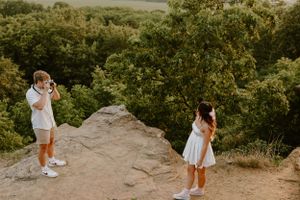 A couple taking photos of each other on a rocky overlook with a forest vista behind them.
