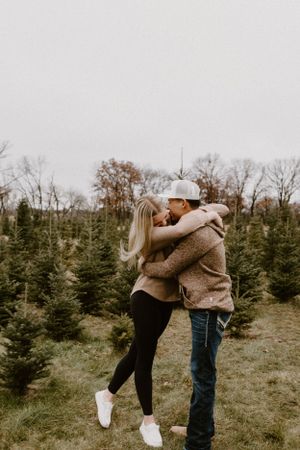 Couple embracing in winter attire at a Christmas tree farm on an overcast day.