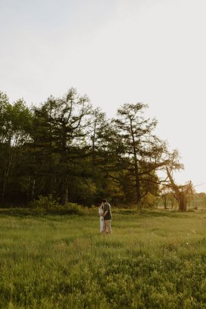 Distant silhouette of couple standing in vast meadow with towering pine trees at dusk.