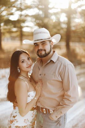 Couple in country western outfits pose together during golden hour in a wooded setting.