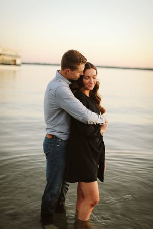 Couple embraces while standing in water at sunset near a peaceful shoreline.