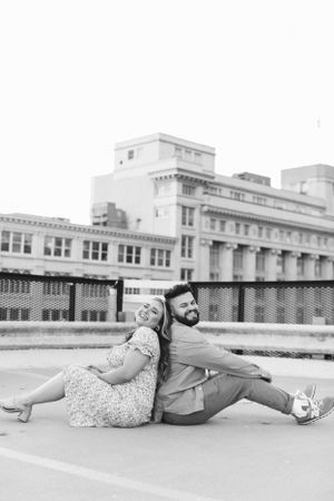 A couple sits back-to-back on a rooftop with urban architecture in the background.
