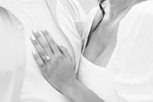 Close-up of a hand wearing a diamond engagement ring against white clothing.
