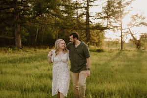 Couple walking together and smiling in a serene meadow with pine trees at sunset.