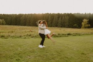 Two figures embrace joyfully while jumping in a grassy field with forest in the background.
