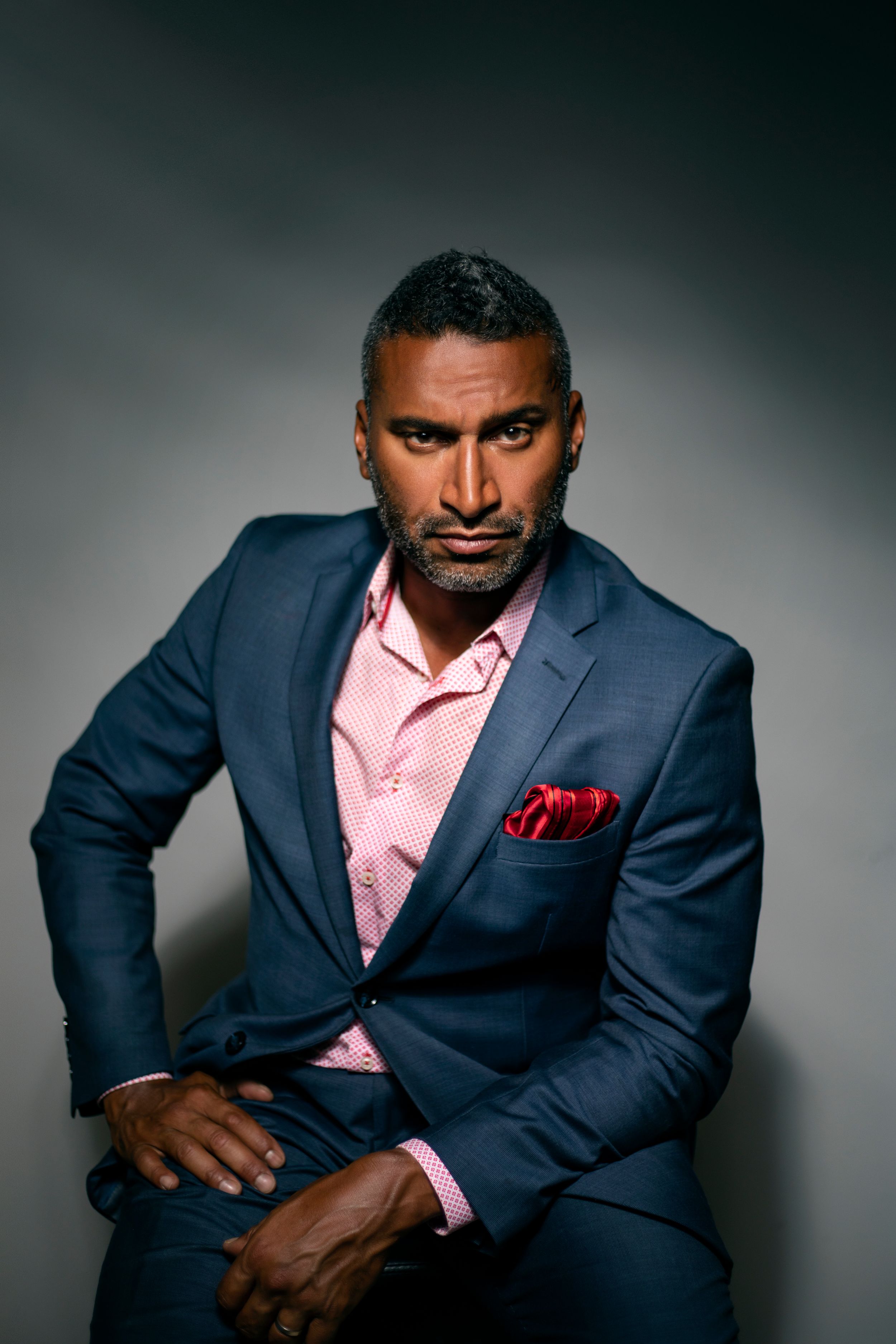 Professional studio portrait of a person wearing a navy suit with pink shirt and red pocket square.