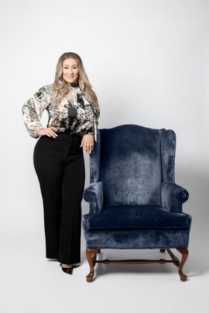 A stylish figure in black pants and patterned blouse poses next to a blue velvet wingback chair against white background.