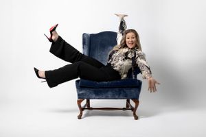Playful pose lounging sideways in a blue velvet armchair wearing patterned top and black pants.