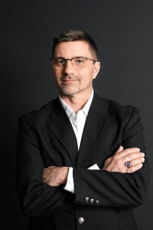 Professional headshot of a person in black suit and glasses against dark background.