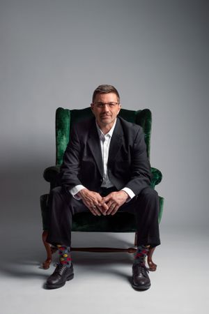 Studio portrait of someone in a black suit seated on an emerald green velvet chair.
