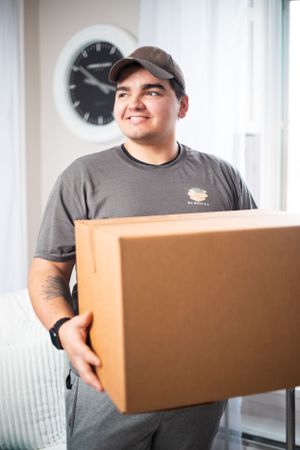 Professional mover carries large brown cardboard box in bright indoor space.