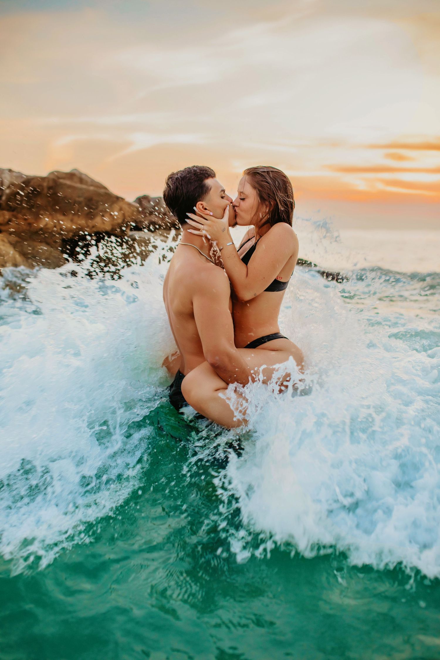 Passionate couple sharing intimate moment in crashing waves at sunset by rocky coastline.