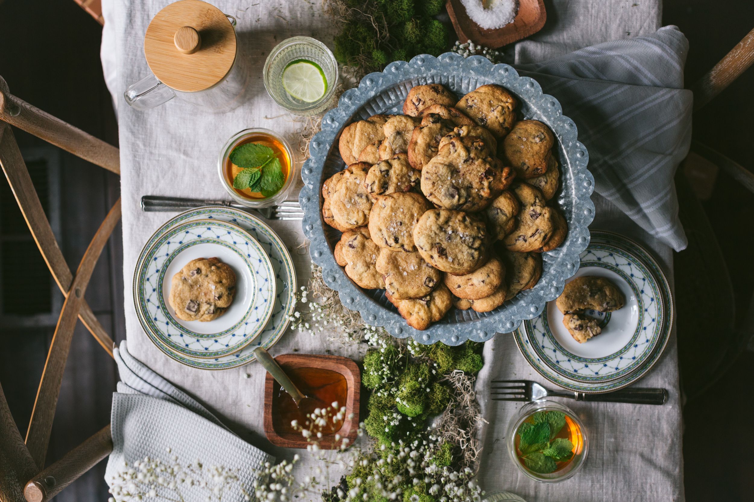 Overhead view of chocolate chip cookies on a decorative plate surrounded by floral arrangements and herbal tea on a rustic table.