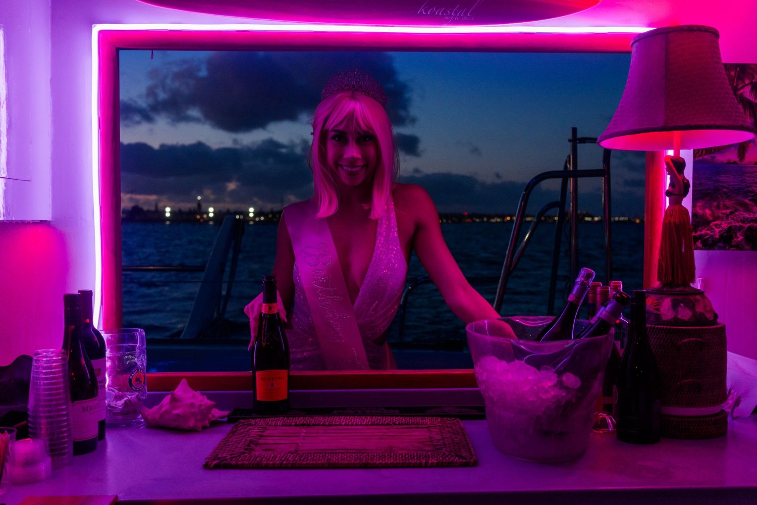 A neon-lit bar scene at night with a purple glow illuminating the bottles and water view through the window.
