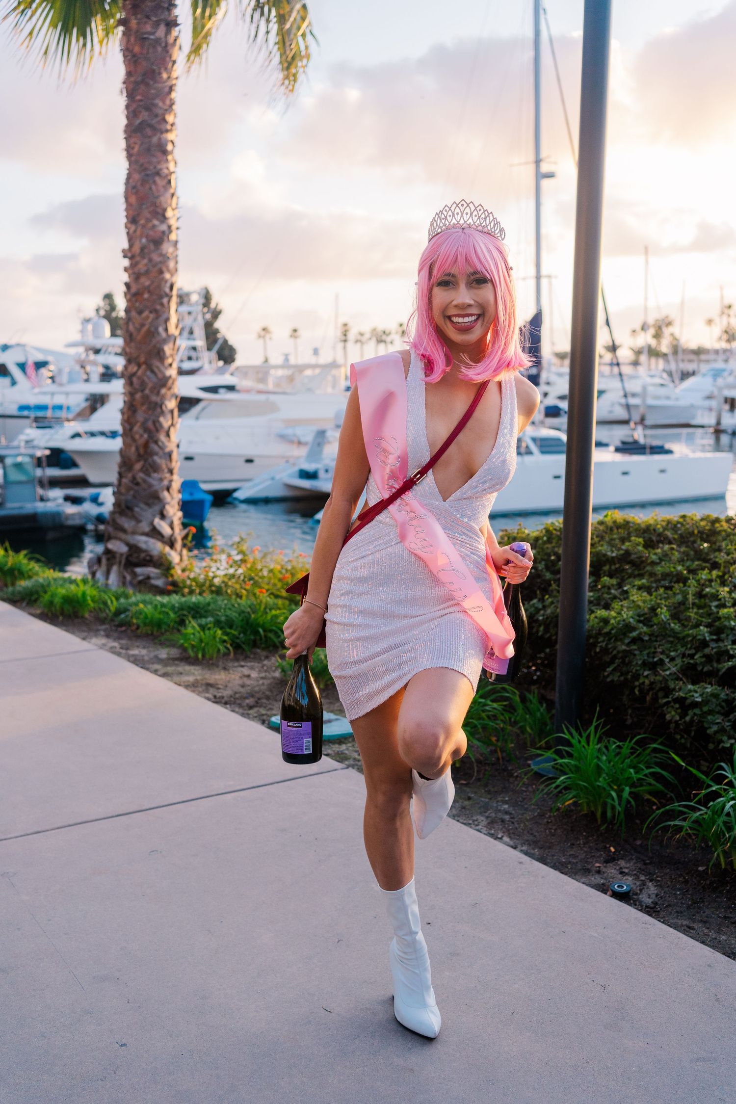 A woman in a white dress and pink wig poses against a marina backdrop with yachts and palm trees at sunset.