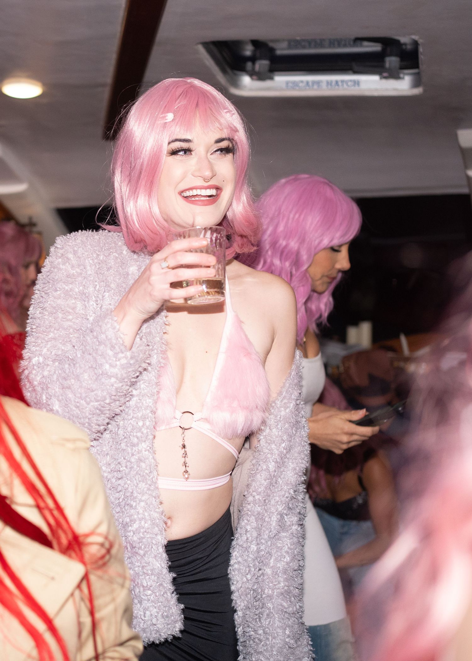 People attend a lively party with pink wigs, sparkly outfits and drinks in a dimly lit room.