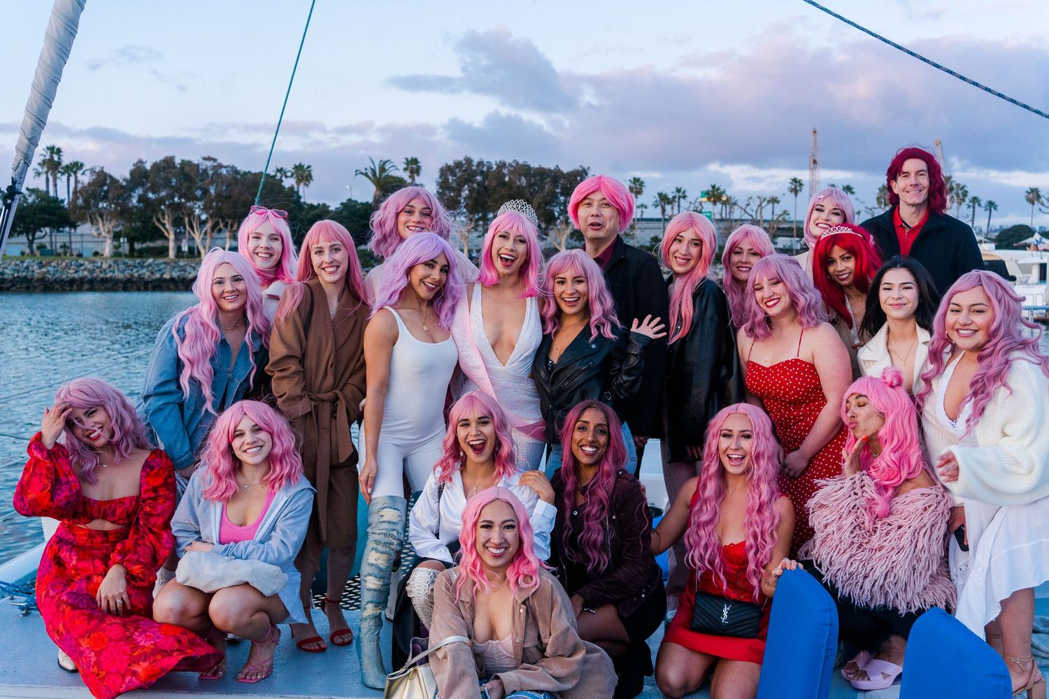 A group wearing pink wigs and festive attire poses together on a boat deck during sunset.