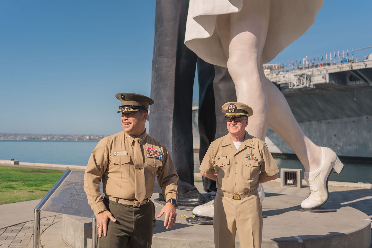 Two military personnel in khaki uniforms stand beside a large ship monument on a sunny day at a waterfront location.